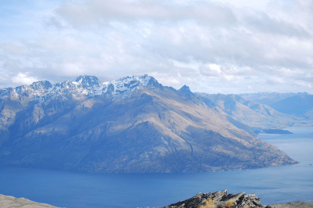 A blue lake surrounds green snow capped mountains. The sky is cloudy with patches of blue