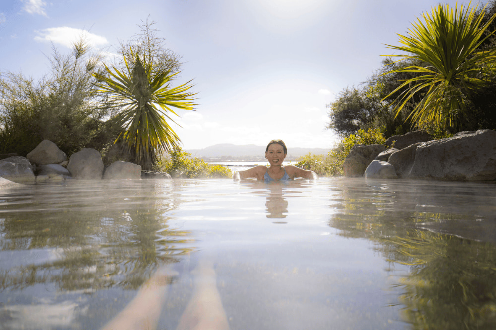 A woman sits in a hot spring on a sunny day. There is plants and scenery in the background