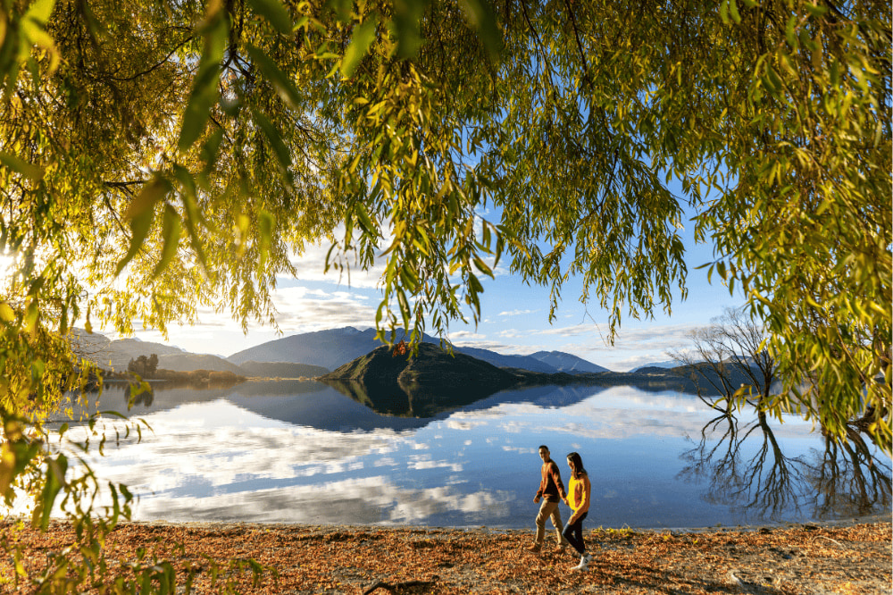 A couple walk along a glassy lake with autumn leaves falling around them. Mountains stand in the background behind the lake