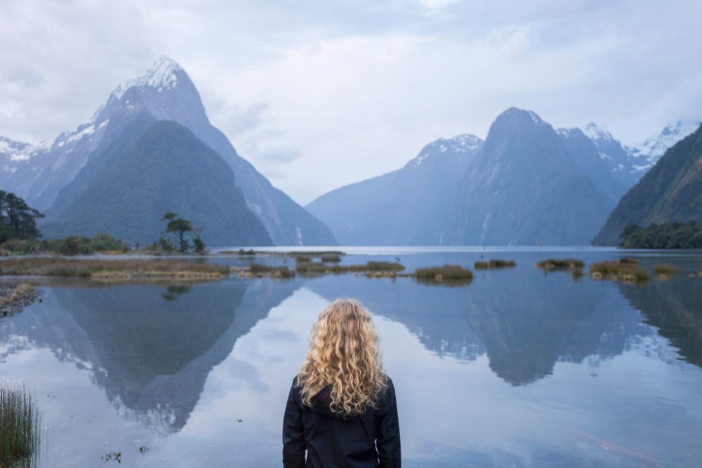 A woman stands with her back to the camera looking out at the vast fiord. The surrounding mountains are reflected in the clear water.