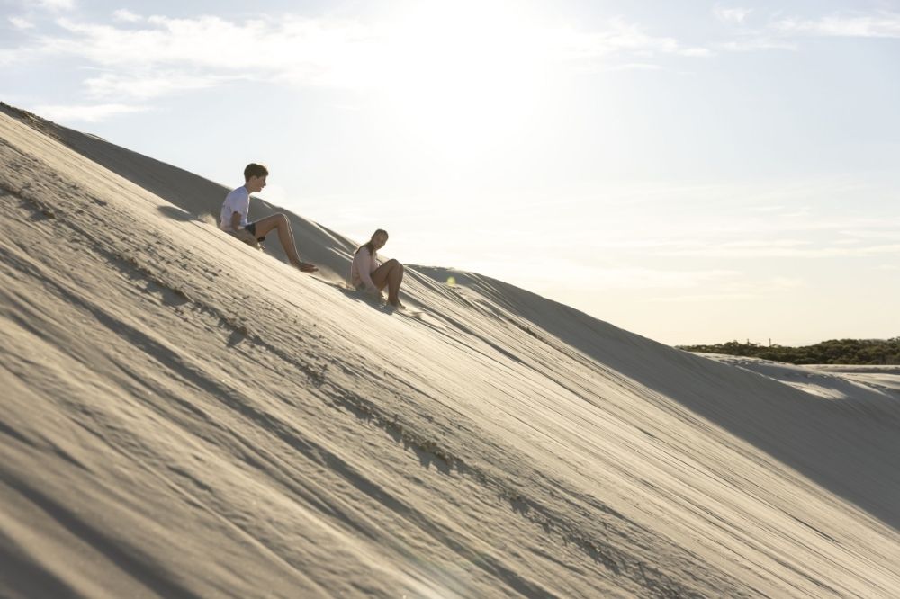 Two people ride down a high sand dune.
