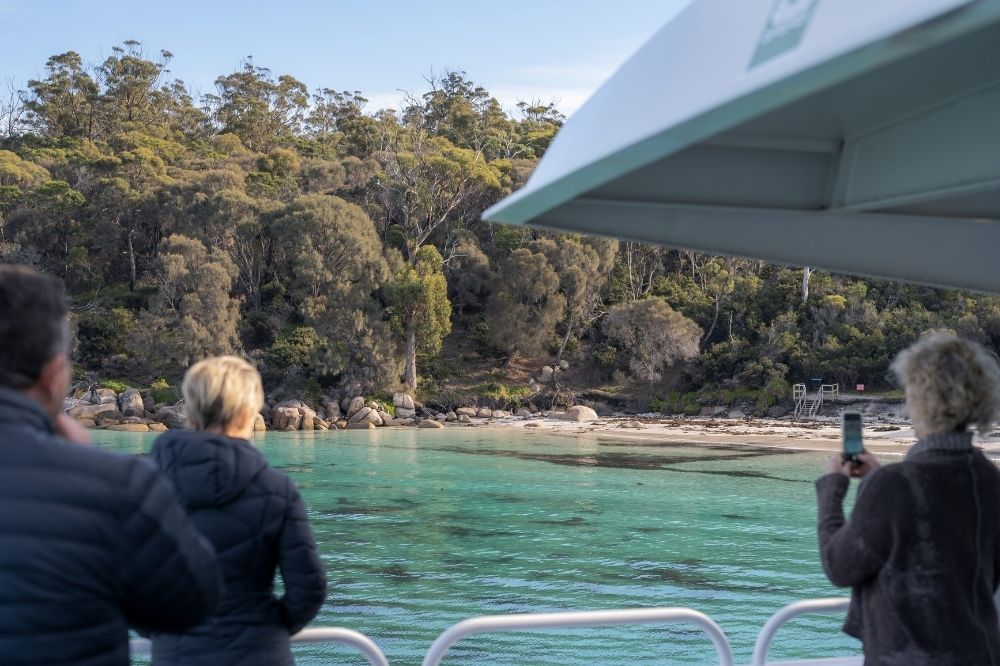 People on a boat look out at a bay with clear blue water. The shore has white sand and trees.