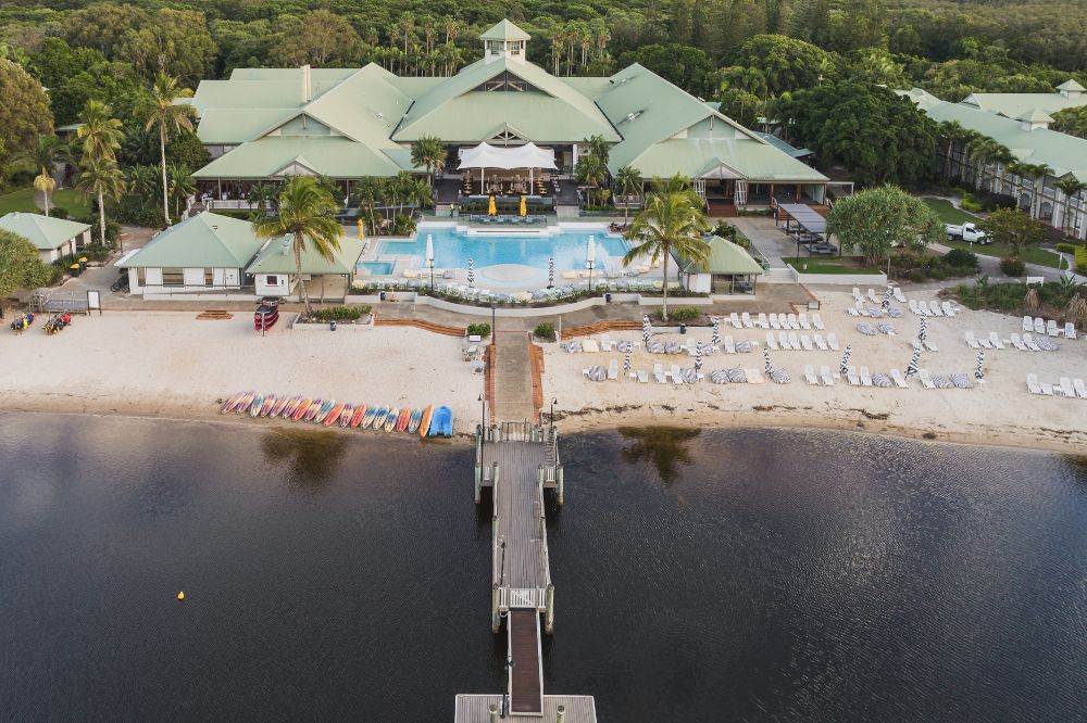 An aerial shot of a resort with a large pool as the centrepiece. There is a boardwalk that does out in to the water.