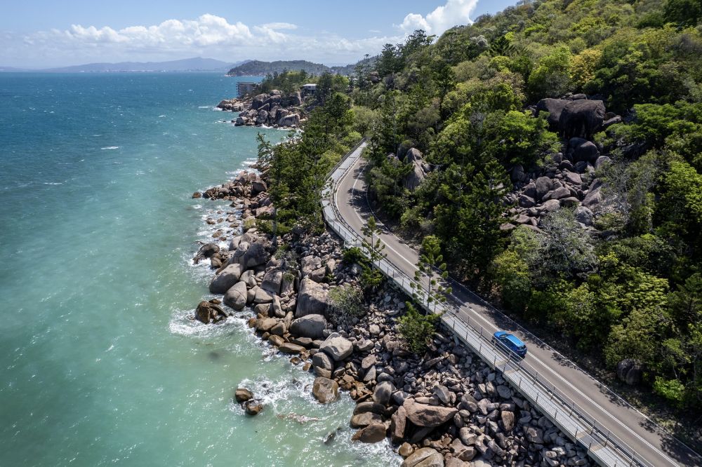A car travels along a coastal road with choppy water in the ocean and rocks and trees on the other side