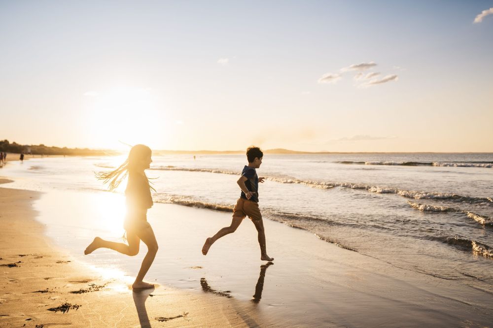 Two kids run towards the ocean. The sun glows on the beach