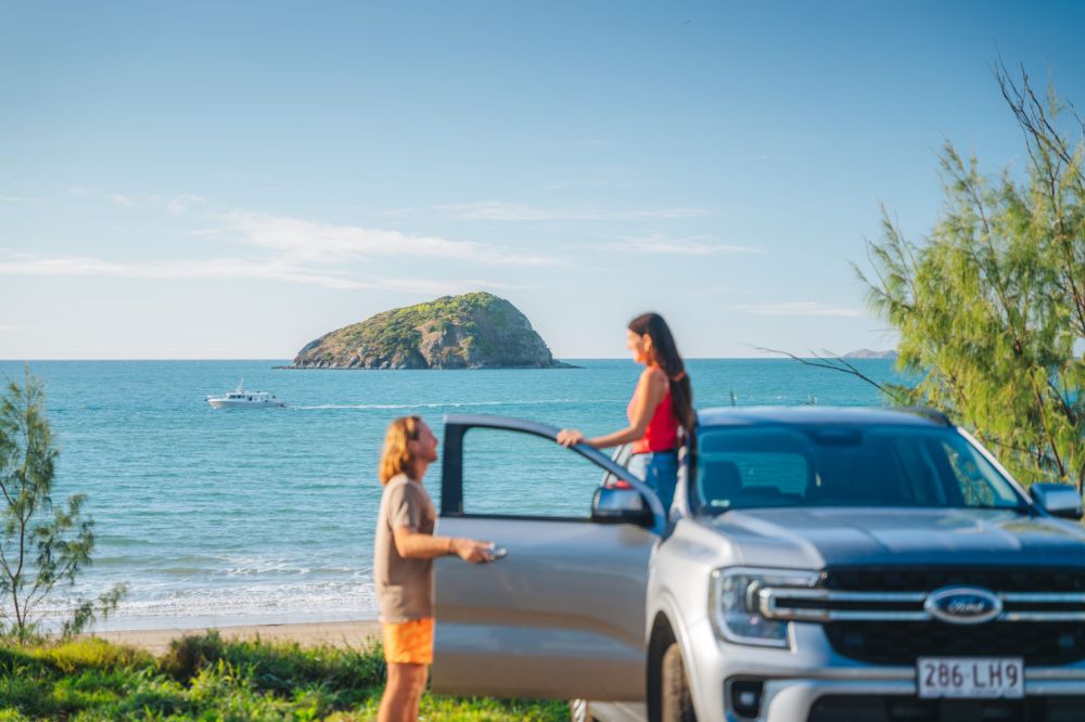 A couple with a car parked near the beach. There's a rocky formation sitting in the ocean and a boat diving past in the water.