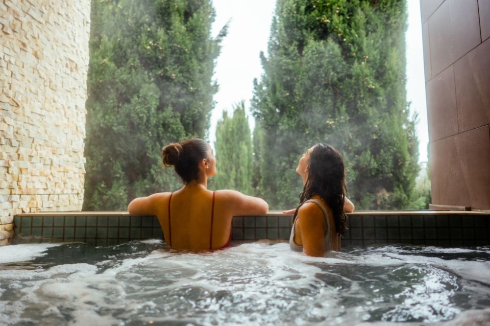 Two people sit in a hot spring looking out at tall green trees.