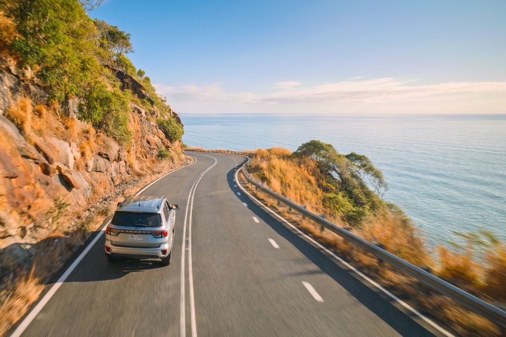 A car drives along a winding road with the calm ocean on the side.
