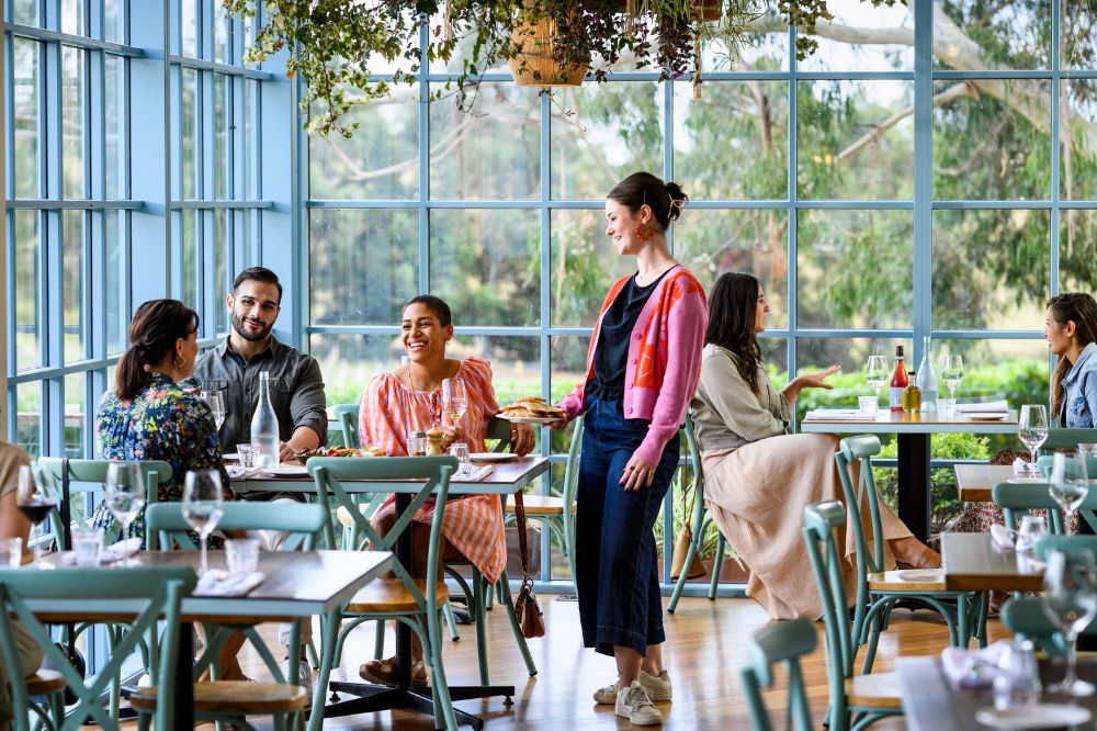 People sit at tables in a winery, the room is lined with windows and greenery is visible outside.