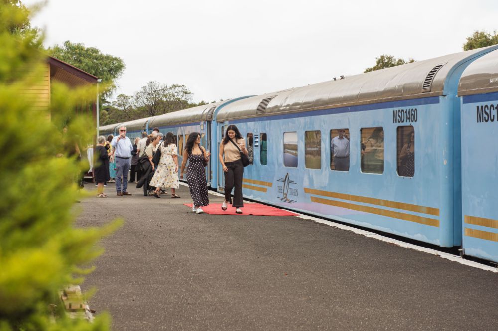 People stand on the platform outside a blue train with glasses of wine in hand