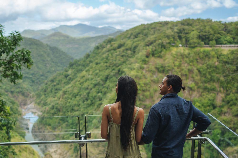 Two people stand at a lookout overlooking tree lined hills and a river below.