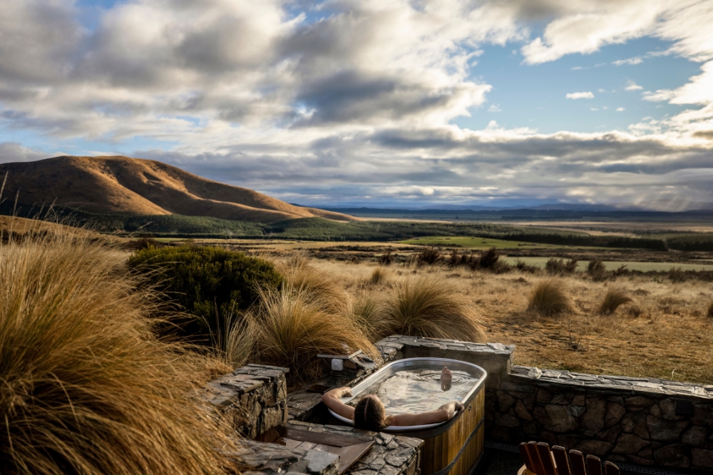 A person soaks in an outdoor bathtub with vast fields in the distance