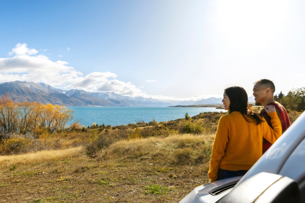 Two people lean on a car looking out on a blue lake with snow capped mountains in the background