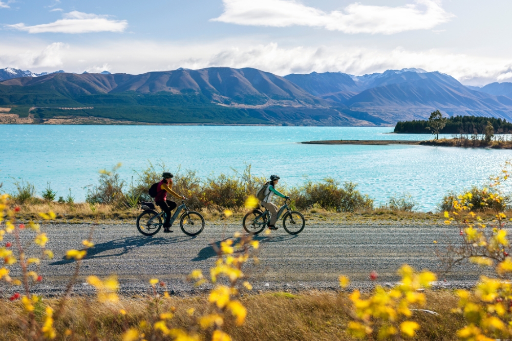 Two people ride bikes along a path next to a lake with bright blue water