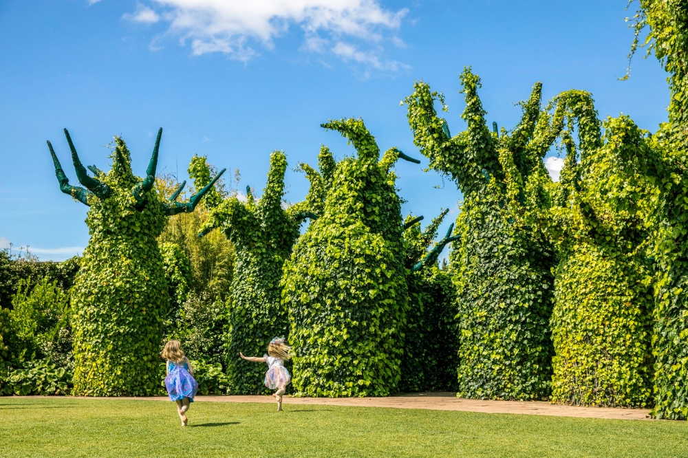 Two children run on the grass with large sculptures covered in ivy surrounding