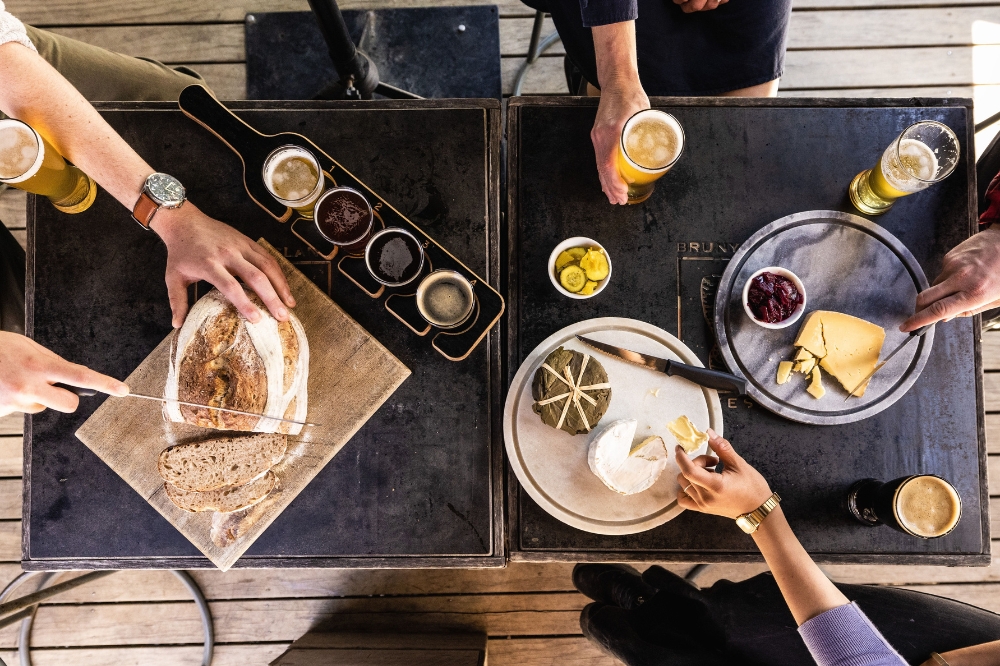 A person slices bread on a table with cheese platters and beers