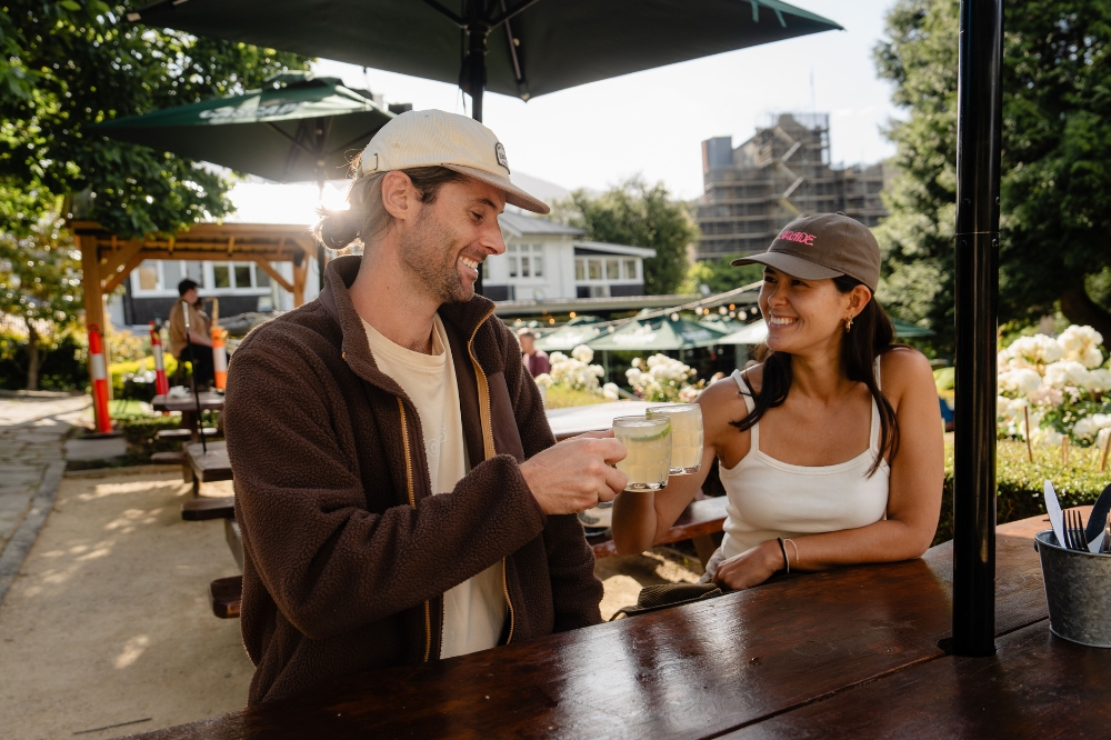 Two people sit under an umbrella with glasses clinking together