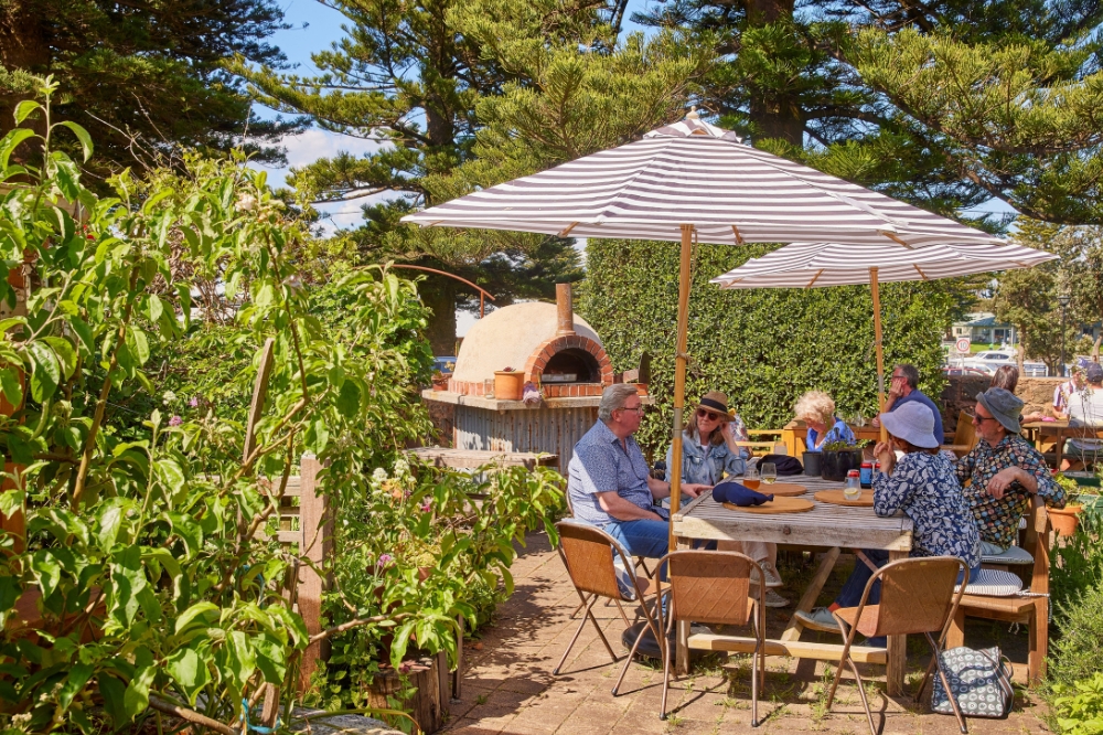 People sit at a table with striped umbrellas above. A woodfire oven is in the background