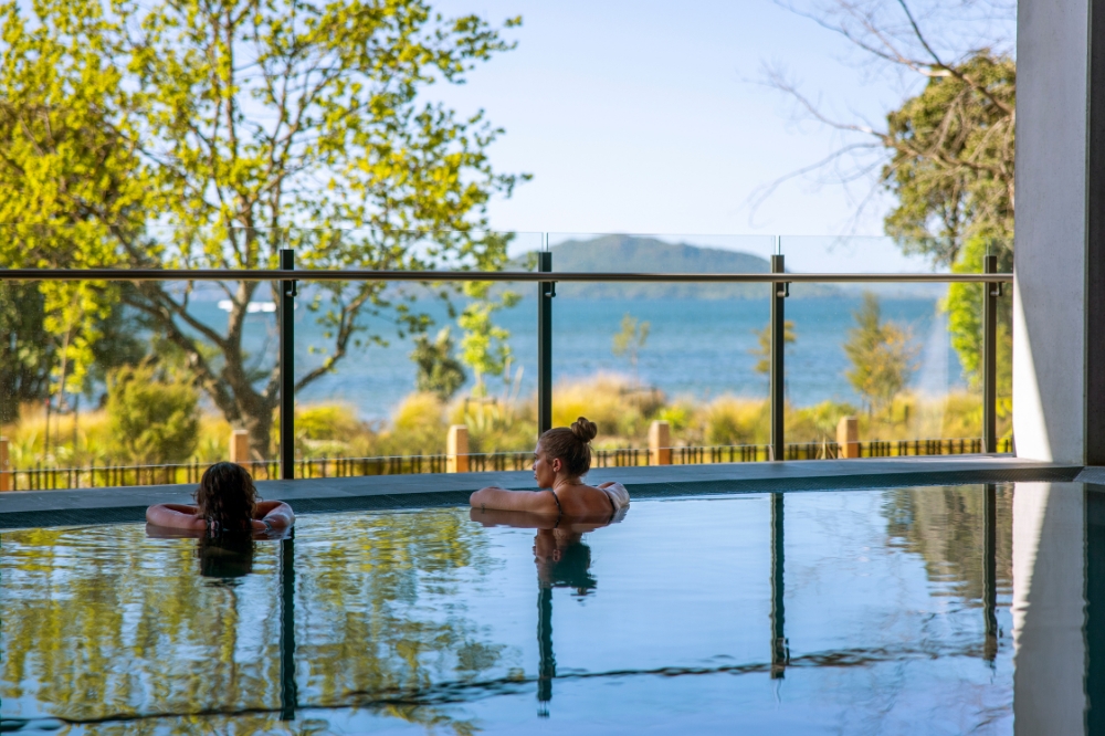 Two people soak in a pool overlooking blue water with trees in the foreground