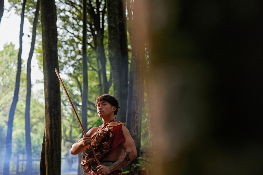 A person dressed in traditional Māori clothes stands among tall trees holding a large piece of wood