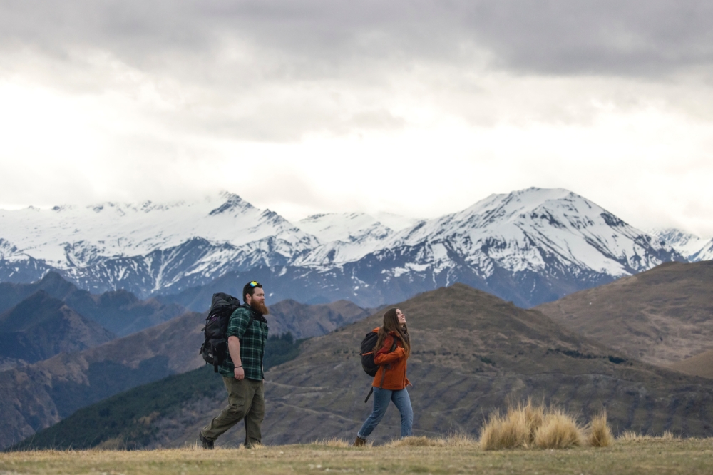 Two people walk along a grassy path looking up at the surrounding snow capped mountains
