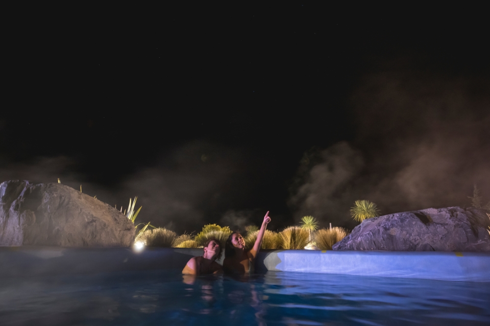 Two people sit in a hot pool looking up at the dark night sky.