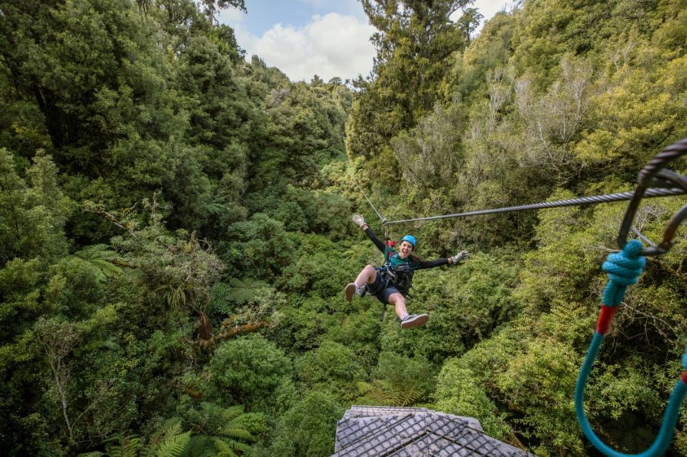 A person smiles at the camera while zip lining over green treetops